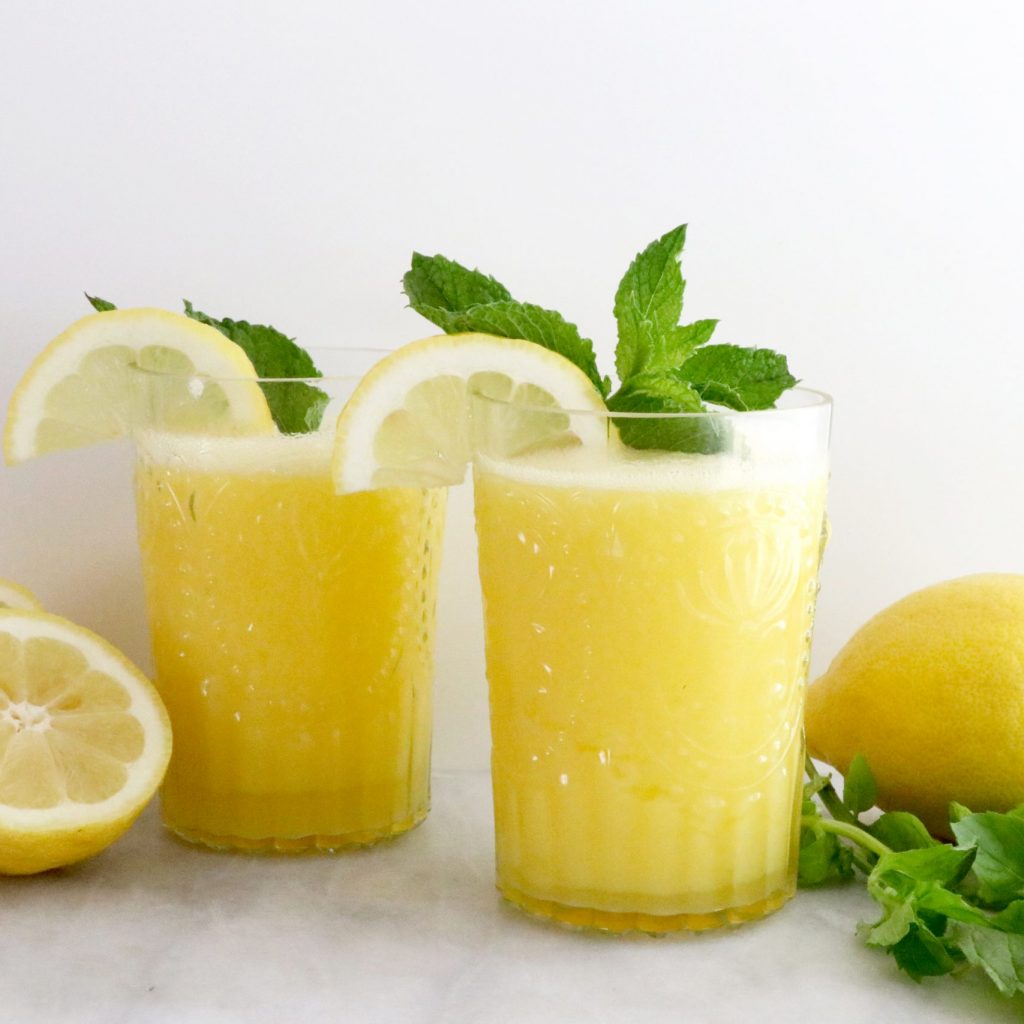 Two glasses full of blended frozen lemonade with a lemon on each side and basil on the right side.